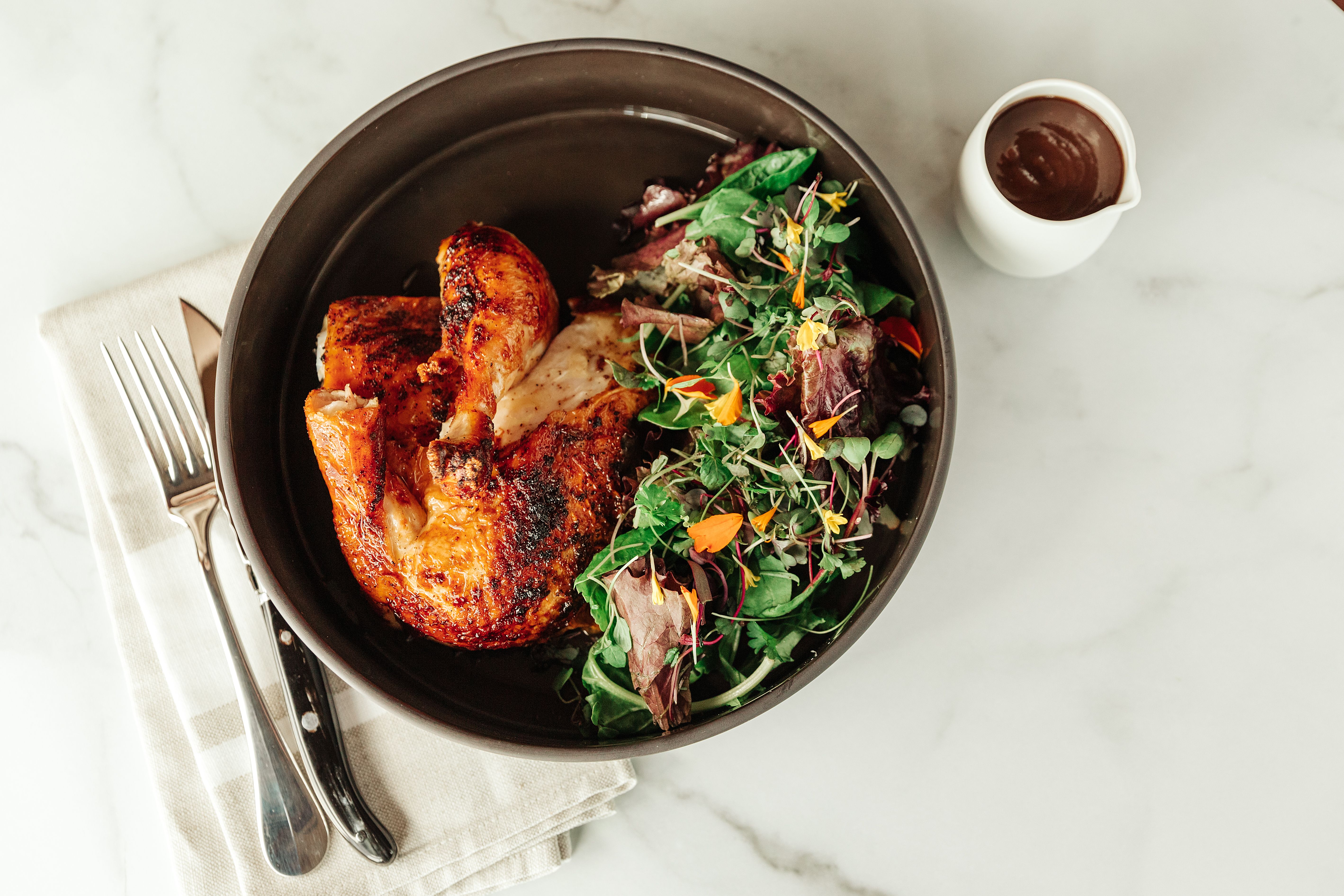 Woodfired chicken dish with microgreen salad in a bowl with silverware on a marble surface.