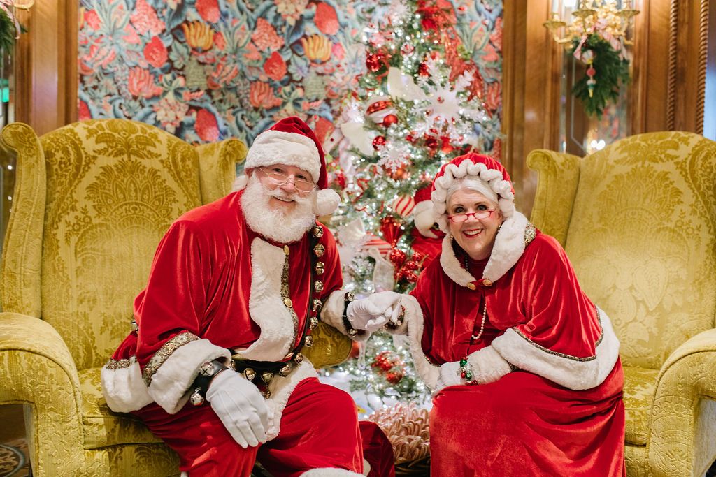 Santa and Mrs. Clause holding hands while sitting in chairs in front of a Christmas tree